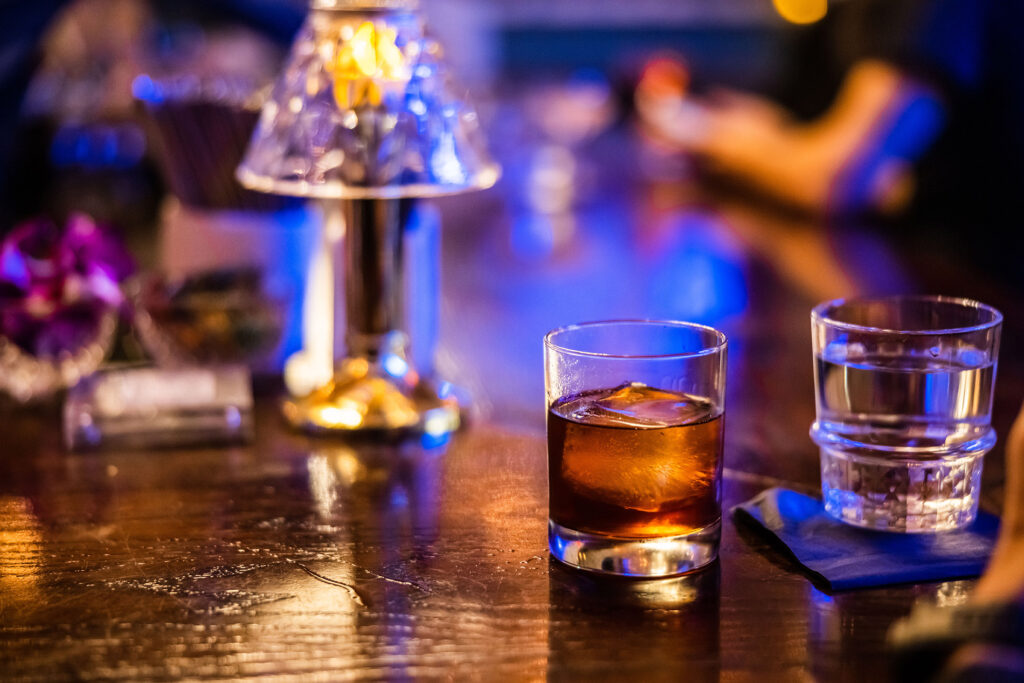 A close up of an old fashioned cocktail with a big ice cube sits on a copper bar top.